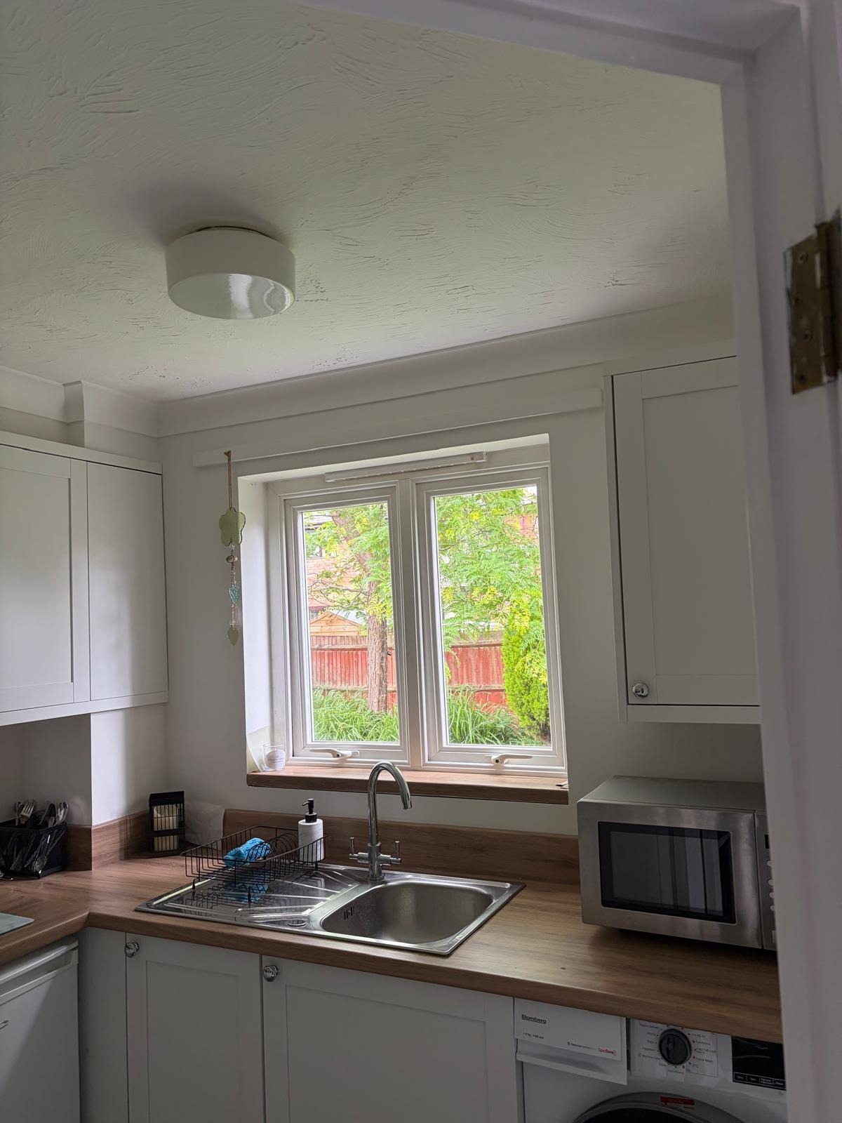 Compact kitchen with a stainless steel sink positioned beneath a double-glazed window overlooking the garden, surrounded by cream cabinetry, wood-effect worktops, and integrated utility appliances.