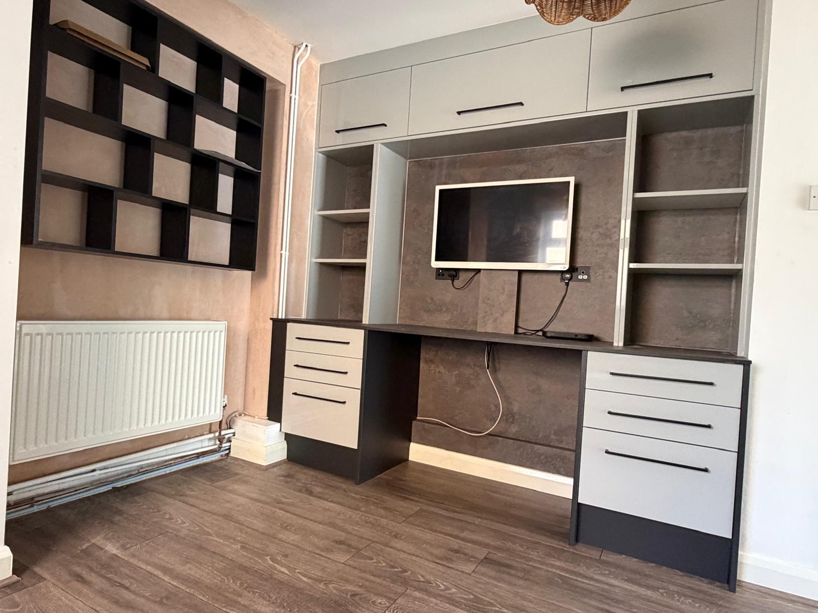 Bespoke fitted home office with grey cabinetry, black worktop, open shelving, wall-mounted TV and matching geometric wall storage.
