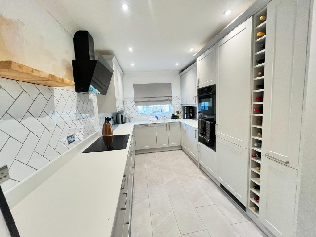 Finished galley kitchen in Fleet featuring pale grey Neves cabinetry, white stone worktops, herringbone tiled splashback, black built-in ovens and an integrated wine rack