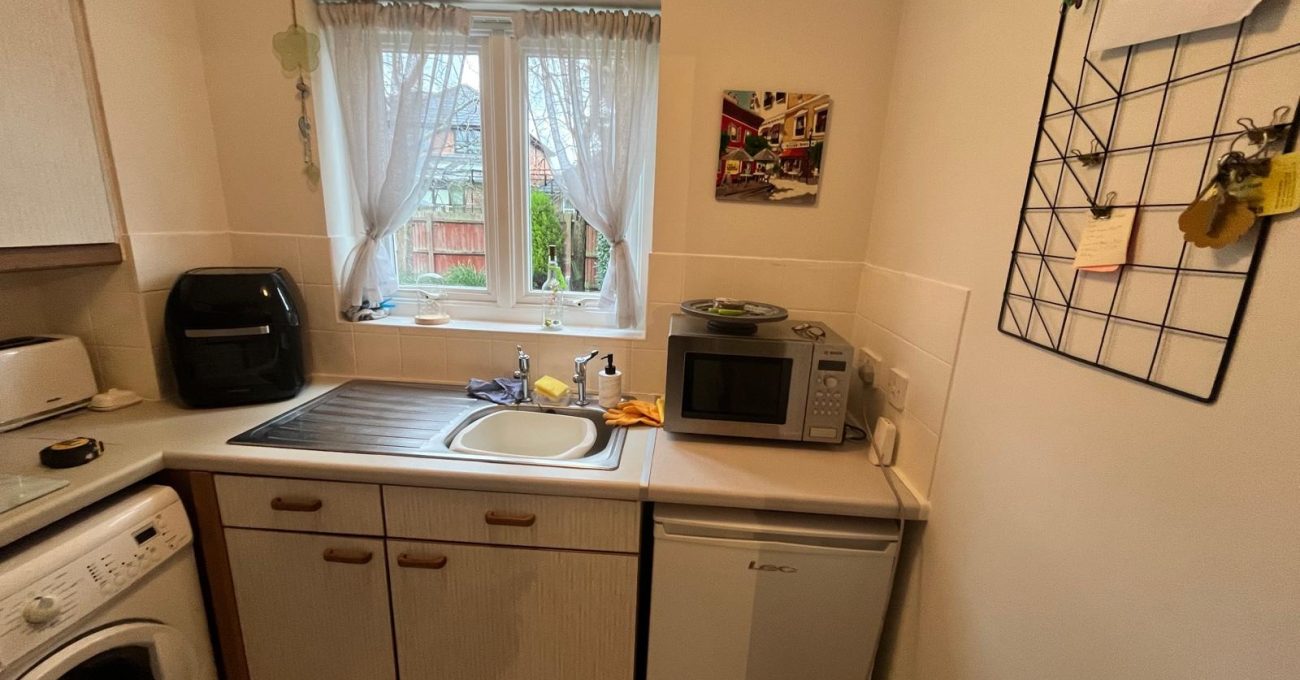 Small kitchen with a window above the sink, cream cabinetry, washing machine, under-counter fridge, microwave, and countertop appliances, creating a functional and well-lit workspace.