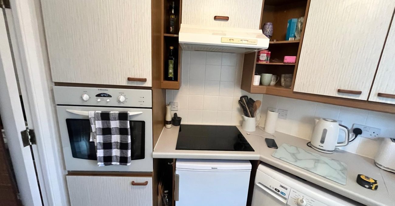 Small fitted kitchen featuring cream wood-effect cabinets, integrated oven, electric hob, extractor hood, under-counter fridge, washing machine, and light-colored worktops in a practical corner layout.