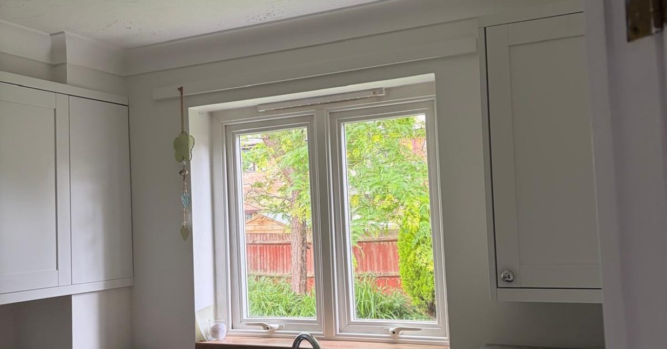 Compact kitchen with a stainless steel sink positioned beneath a double-glazed window overlooking the garden, surrounded by cream cabinetry, wood-effect worktops, and integrated utility appliances.