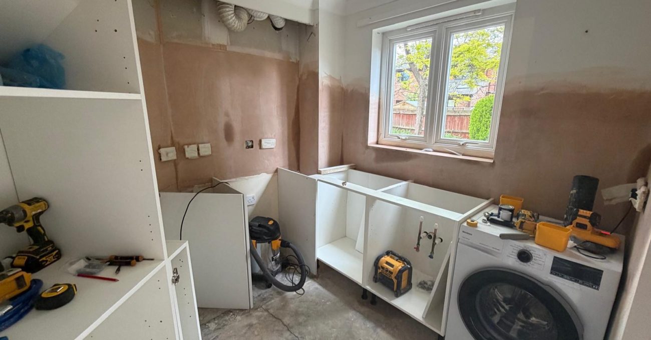 Partially completed kitchen installation featuring shaker-style cabinet carcasses, utility connections, washing machine, exposed wall preparation, and construction tools during the fitting process.