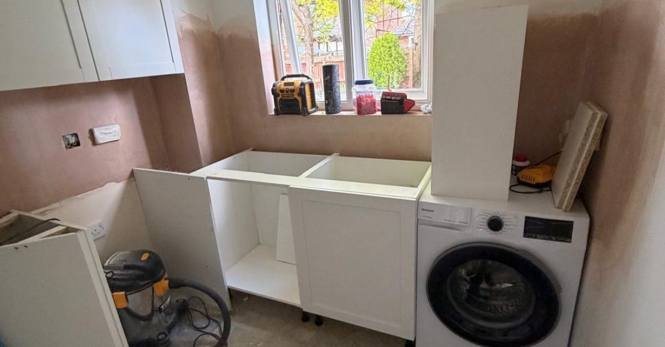 Compact kitchen renovation showing newly installed base cabinets, washing machine, window-facing sink area, unfinished worktops, and preparation work underway before final fitting and finishing.