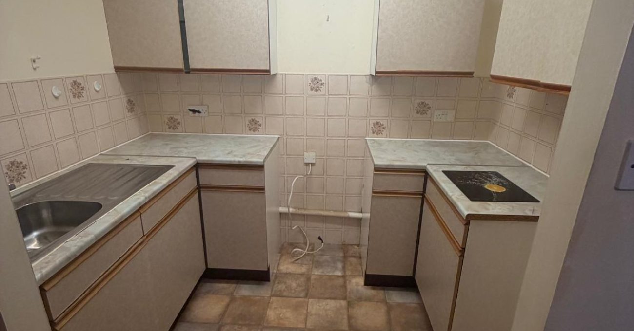 Wide view of the dated U-shaped kitchen layout with beige cabinetry, patterned wall tiles and terracotta-style flooring before renovation