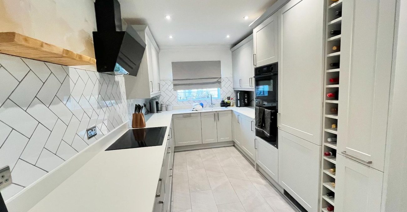 Finished galley kitchen in Fleet featuring pale grey Neves cabinetry, white stone worktops, herringbone tiled splashback, black built-in ovens and an integrated wine rack