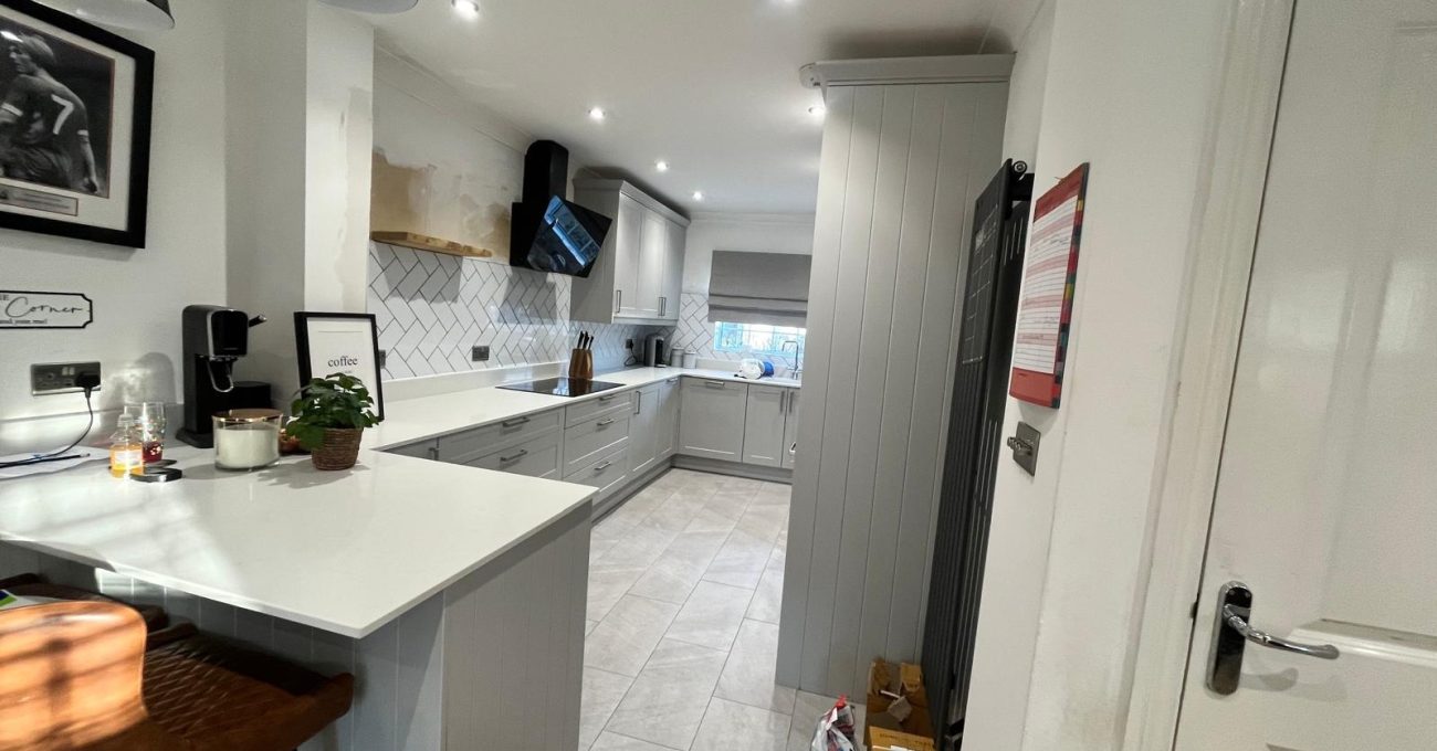 View from the breakfast bar of a completed Fleet kitchen showing white stone countertops, grey cabinetry, pendant lighting and new tiled flooring leading to the cooking area