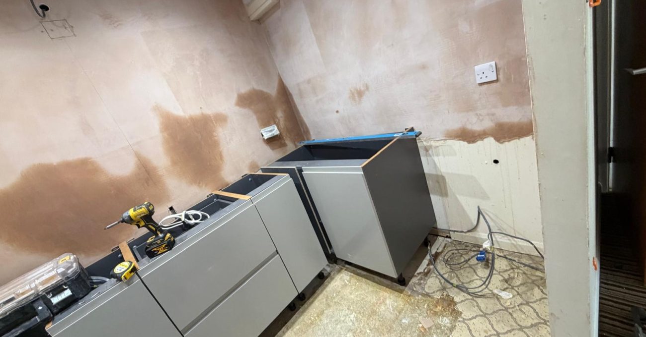 New grey kitchen base units fitted along stripped-back walls during the SJM Kitchens installation, with power tools and materials on site