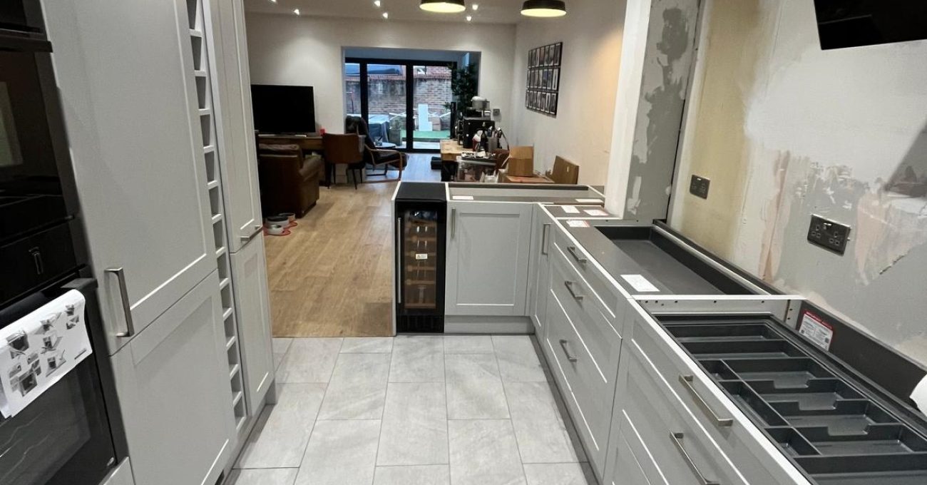 Galley kitchen in Fleet mid-installation showing grey Neves cabinetry, integrated wine cooler, new floor tiles and a view through to the open-plan living area